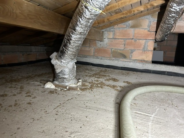 Combles perdus avant travaux : la dalle est nue et poussiéreuse, une gaine de ventilation descend le long du mur de briques.